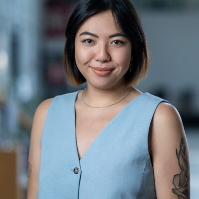 Bild von Sally Chan. Sie trägt eine hellblaue Bluse, eine silberne Kette und einen Nasenring. Auf dem linken Arm ist ein abstraktes Tattoo.